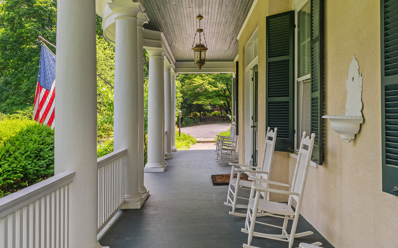 A spacious front porch with white columns, rocking chairs, and a hanging lantern. Green shutters frame the windows, while an American flag marks the side—a-list: June 2026 style—lush greenery lining the view beyond the railing.