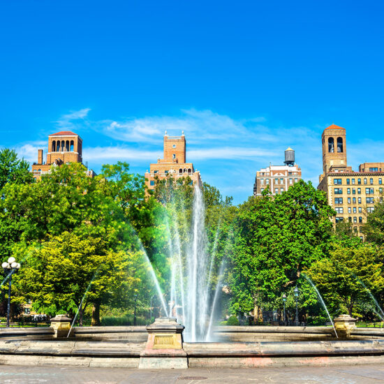 A large fountain sprays water upwards in the center of a park, surrounded by green trees. Behind the trees, historic buildings with red rooftops evoke classic NYC residential rental charm against a clear blue sky.
