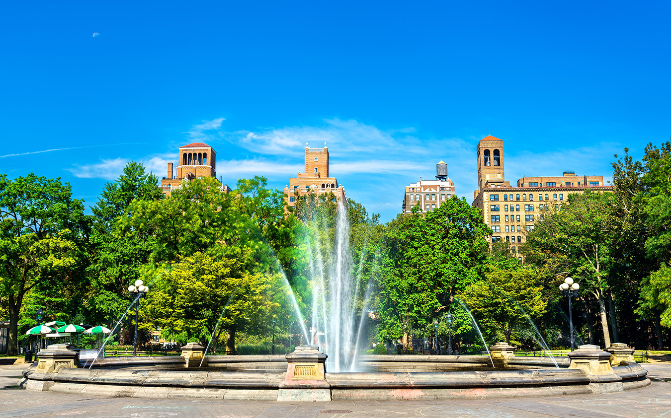 A large fountain sprays water upwards in the center of a park, surrounded by green trees. Behind the trees, historic buildings with red rooftops evoke classic NYC residential rental charm against a clear blue sky.