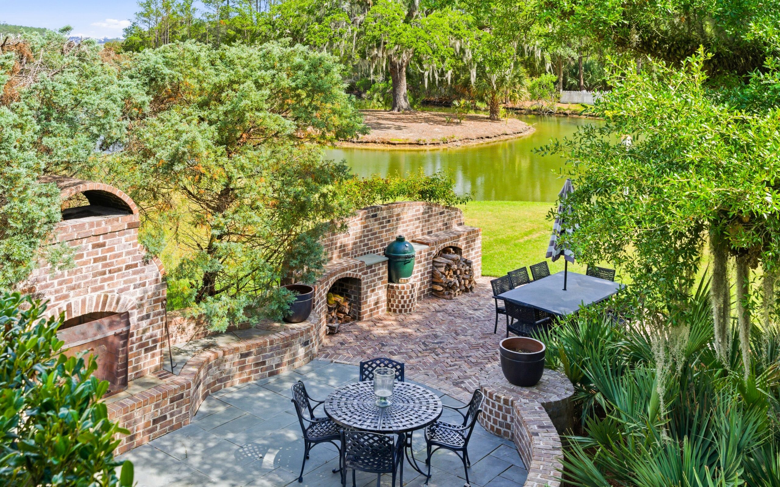 Brick patio with outdoor kitchen, grill, and oven overlooks a small pond. Black metal dining tables and chairs are nestled in lush greenery and trees, creating a peaceful backyard garden setting.