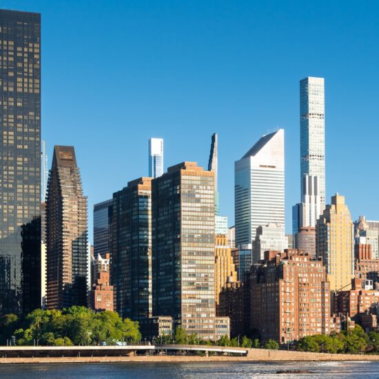A clear, sunny view of the New York City skyline featuring modern and classic skyscrapers hints at the thriving Manhattan luxury sales market, with trees and water in the foreground.