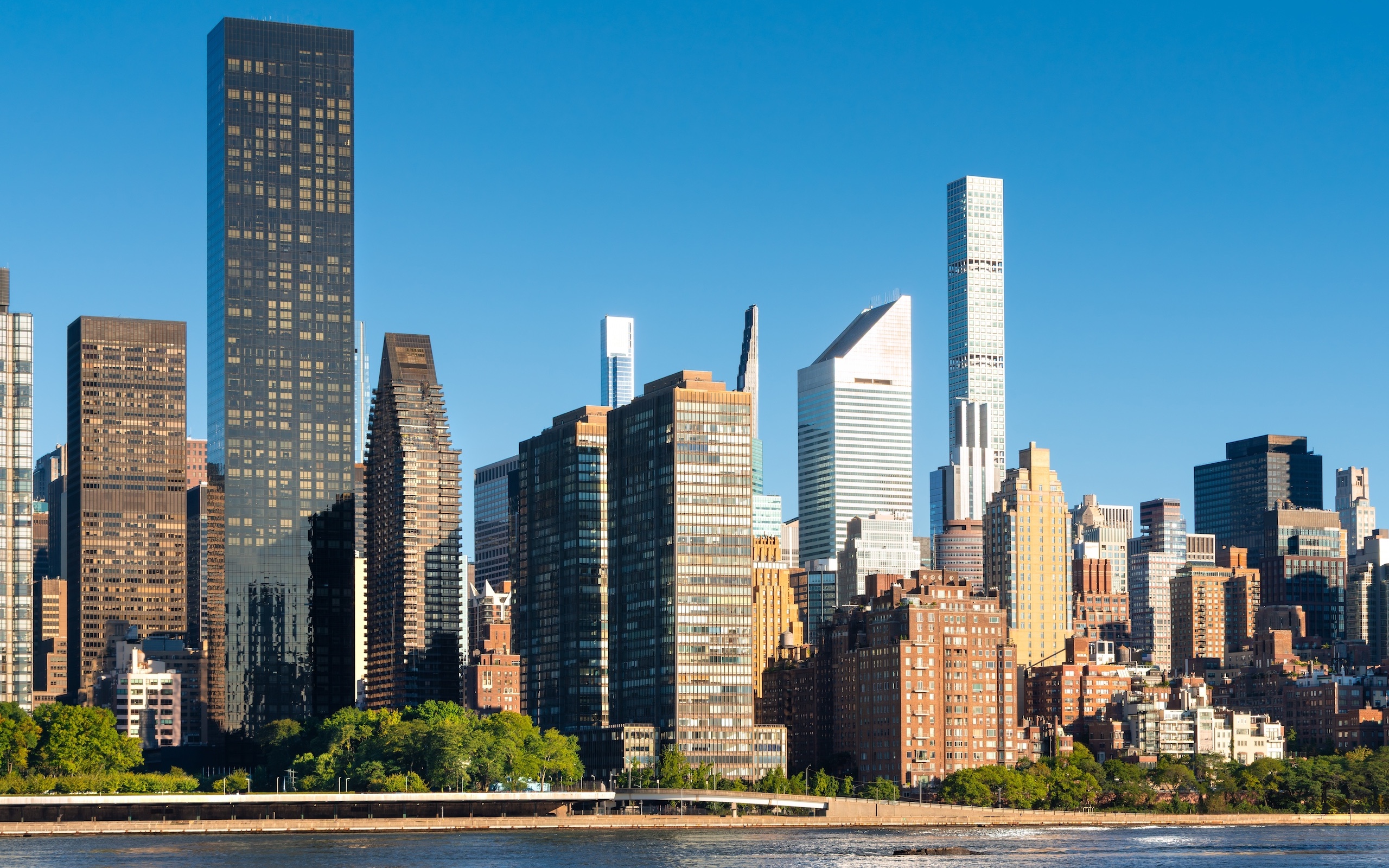 A clear, sunny view of the New York City skyline featuring modern and classic skyscrapers hints at the thriving Manhattan luxury sales market, with trees and water in the foreground.