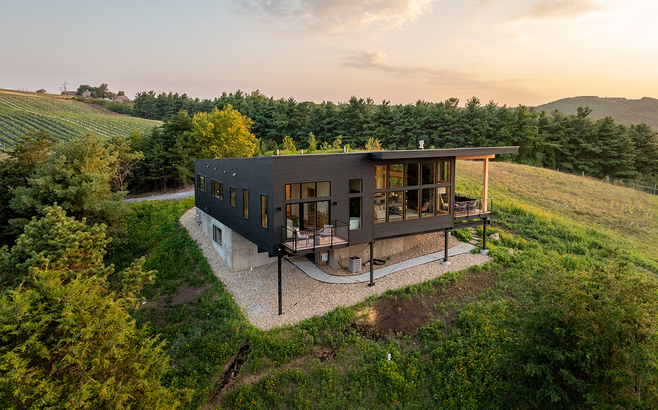 Modern black rectangular house with large windows and a slanted roof, surrounded by greenery, trees, and hills, under a partly cloudy sky at sunset.