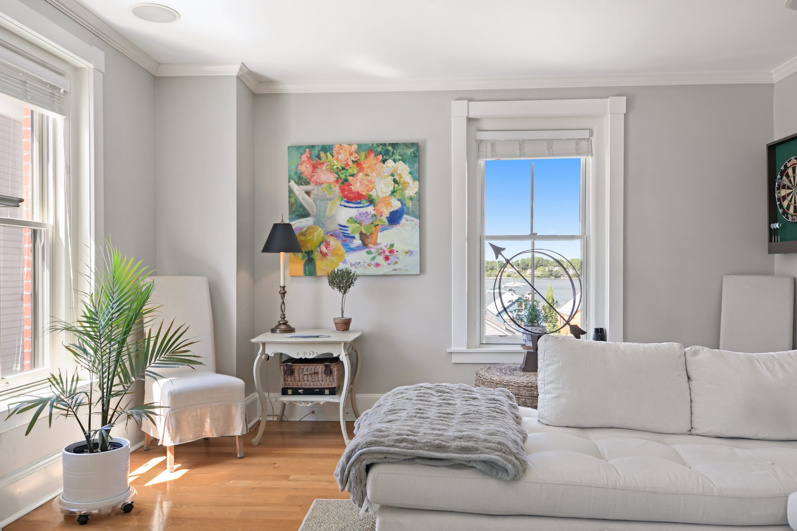 Bright living room with a white sofa, potted plant, side table with lamp, colorful floral painting on the wall, window with outdoor view, and decorative items on the table and windowsill—perfectly styled for an A-list gathering in August 2026.