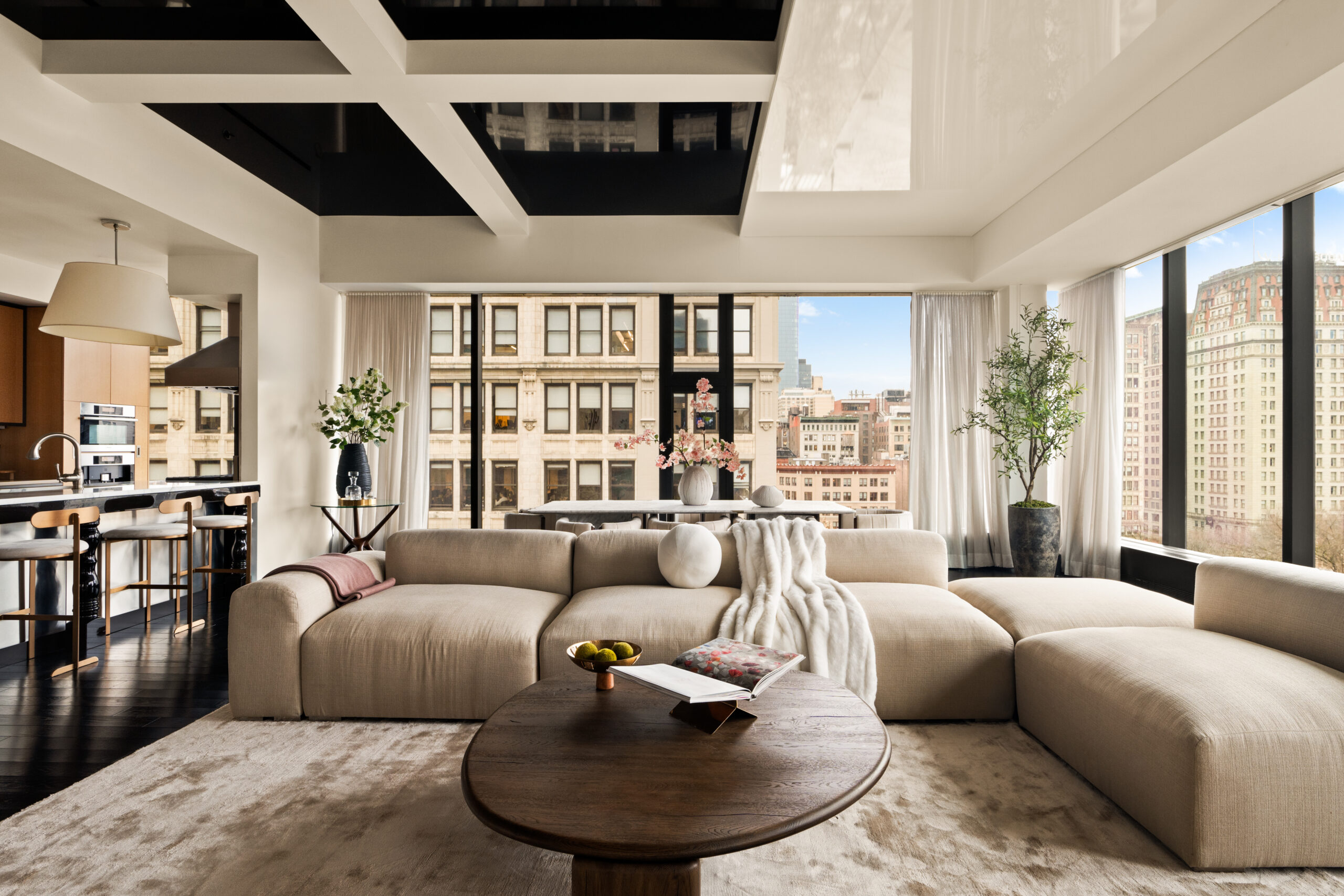 Spacious modern living room with a large beige sectional sofa, round wooden coffee table, and floor-to-ceiling windows overlooking a cityscape—a setting worthy of the a-list: August 2026. Decor includes plants, flowers, and a cozy throw blanket.