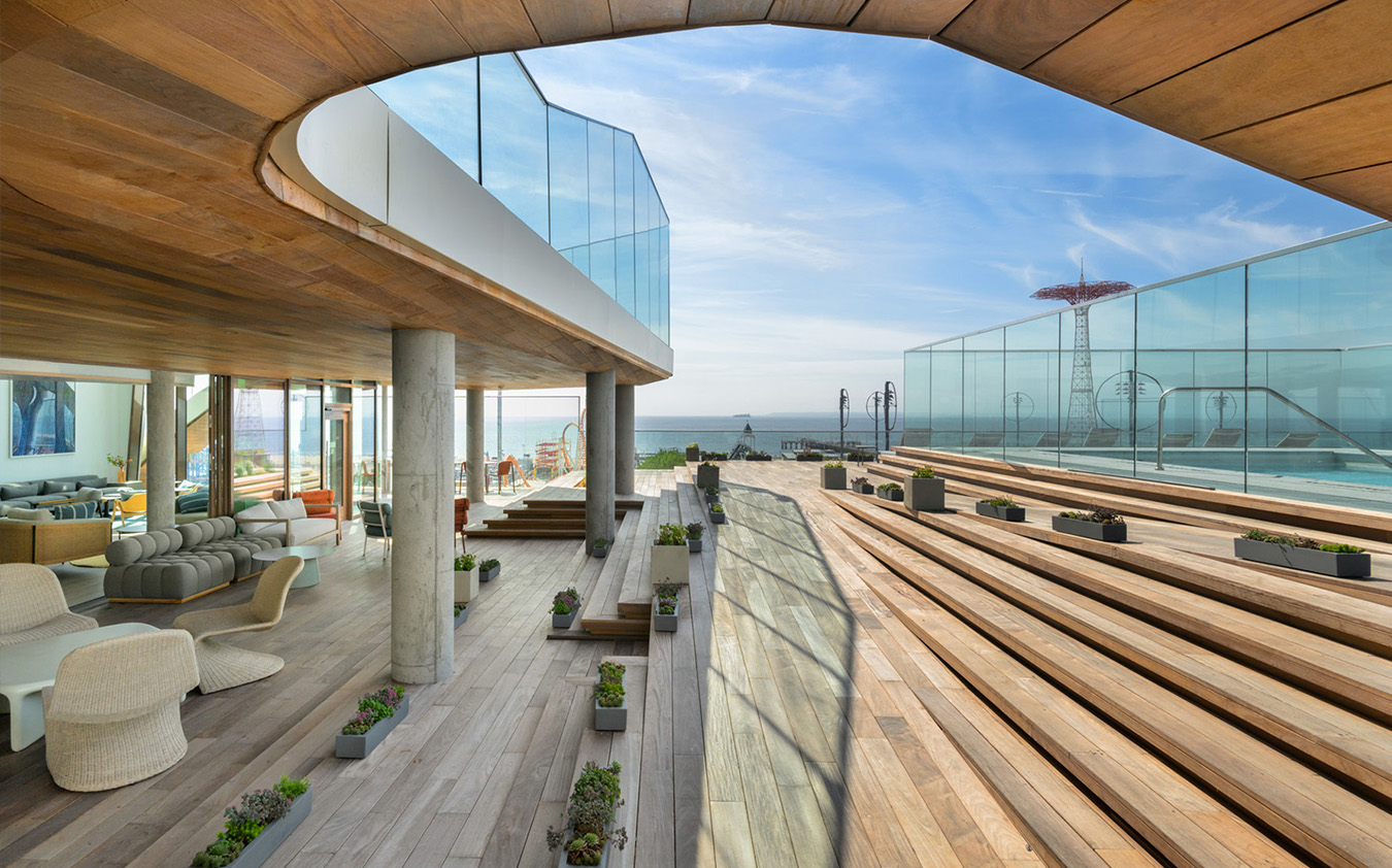 Modern rooftop terrace with wooden decking, tiered seating, glass railings, potted plants, and an outdoor seating area. The terrace overlooks the ocean with a clear blue sky and a red tower visible in the distance.