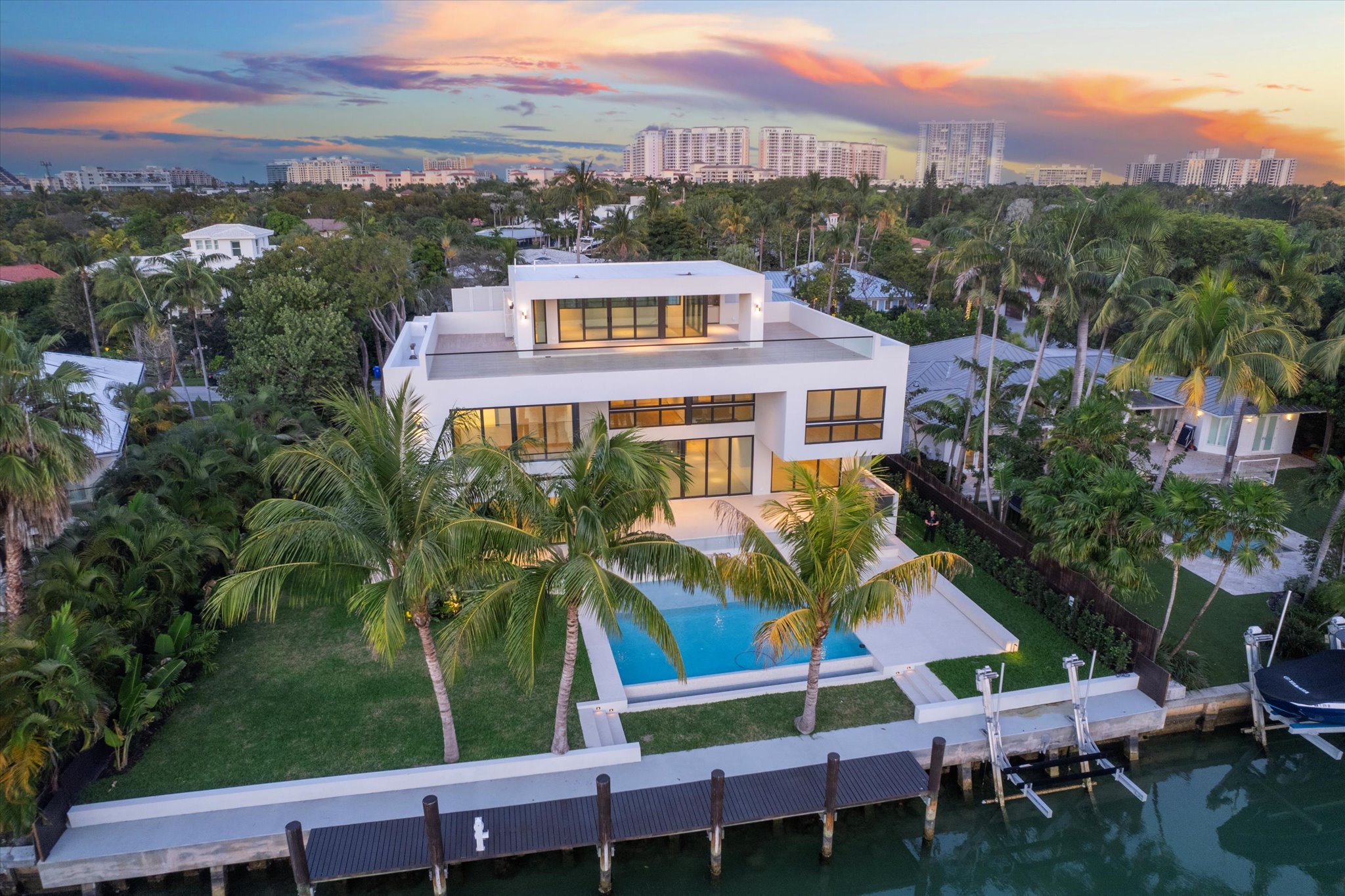 Modern three-story waterfront house with large windows, swimming pool, palm trees, and private dock, set against a sunset sky with city buildings in the distance—a true A-list retreat perfect for August 2026.