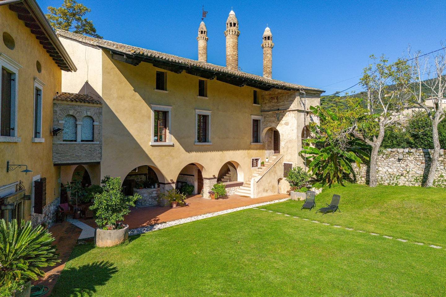 Historic villa with arched walkways, stone chimneys, and yellow stucco walls, surrounded by lush green lawn, potted plants, and outdoor lounge chairs under a clear blue sky—a perfect A-list retreat for August 2026.