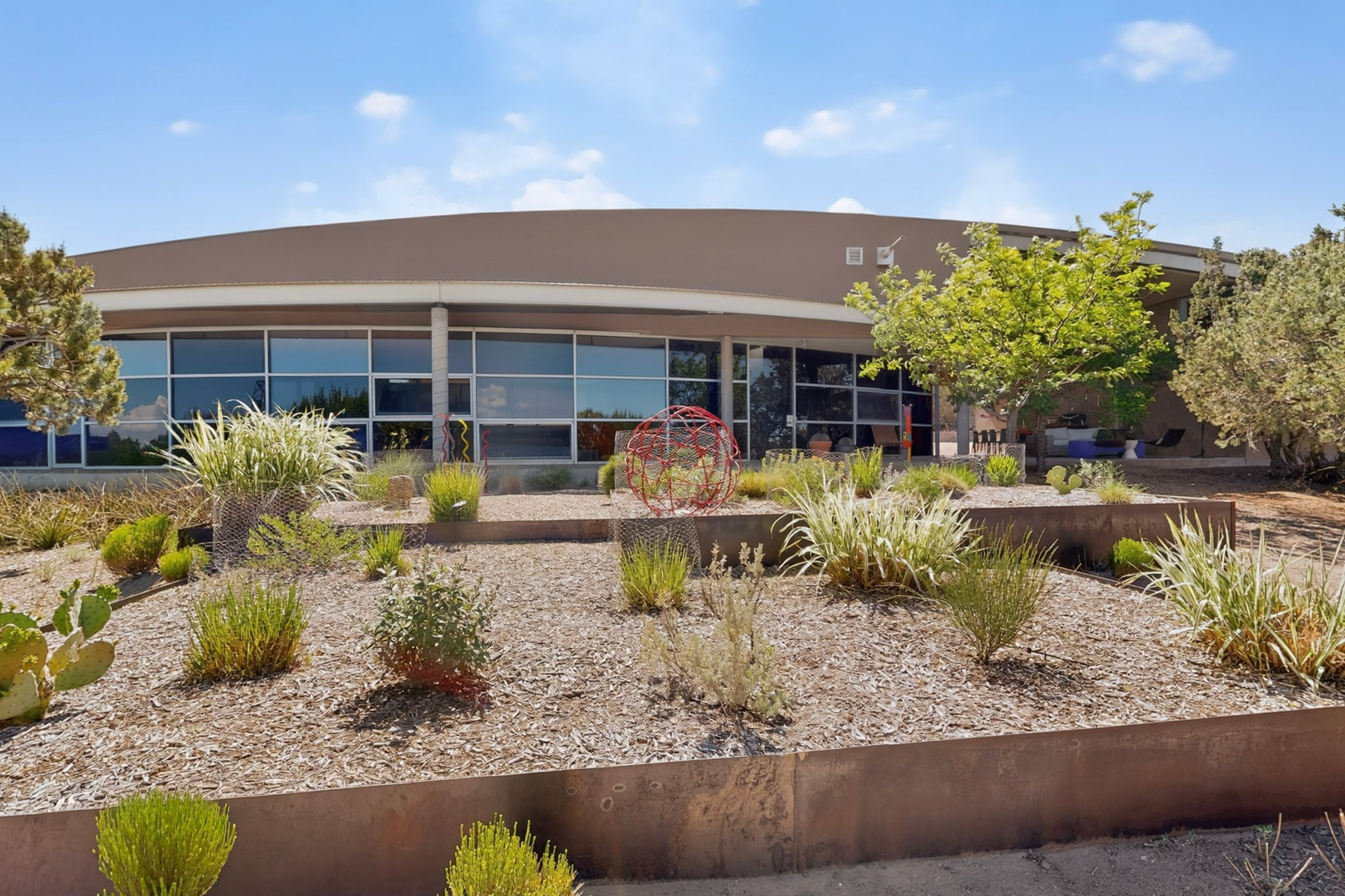 Modern building with large windows, surrounded by desert landscaping, various shrubs, and small trees. A striking red metal sculpture stands in the center of the foreground garden. Blue sky with scattered clouds above adds to the serene setting—an a-list architectural destination poised to impress in August 2026.