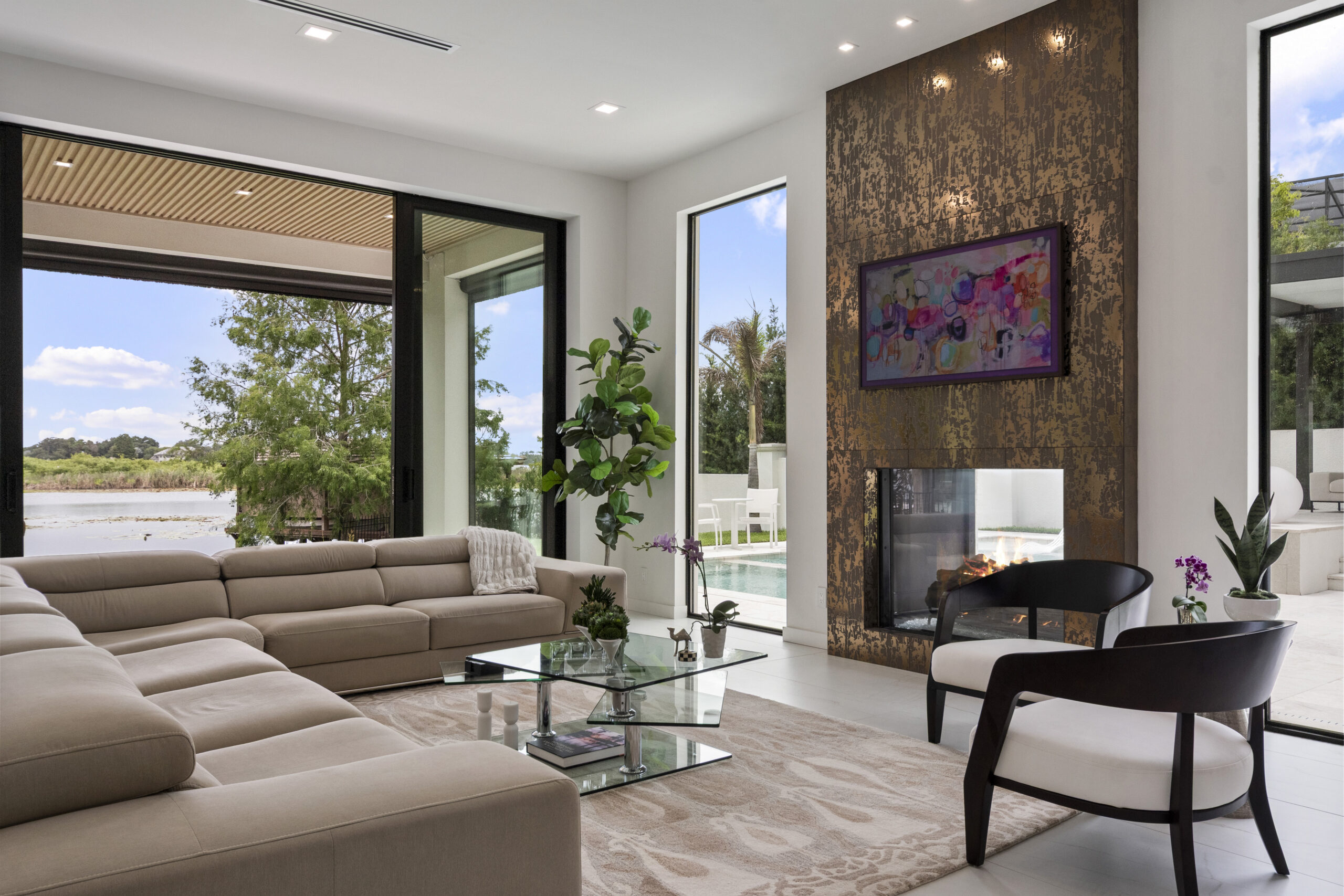 Modern living room with large beige sectional sofa, glass coffee table, fireplace with textured gold wall, colorful art, indoor plants, and floor-to-ceiling windows overlooking a pool and outdoor scenery—a true a-list space ready for August 2026 gatherings.