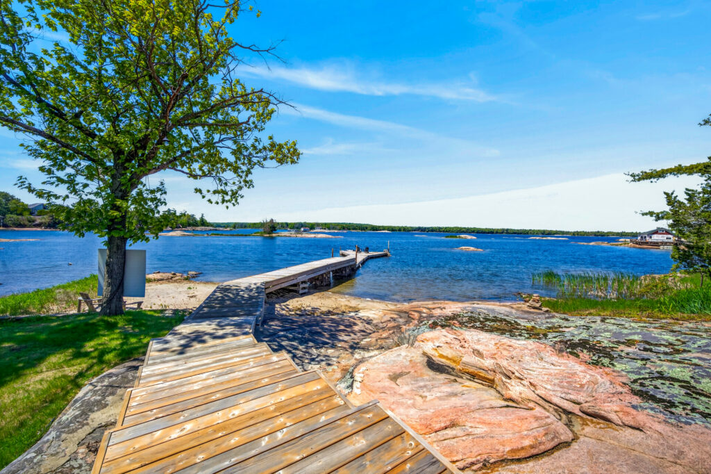 A wooden boardwalk leads to a dock over clear blue water, showcasing inviting summer amenities amid rocks, grassy areas, and a tree in the foreground under a bright, sunny sky.