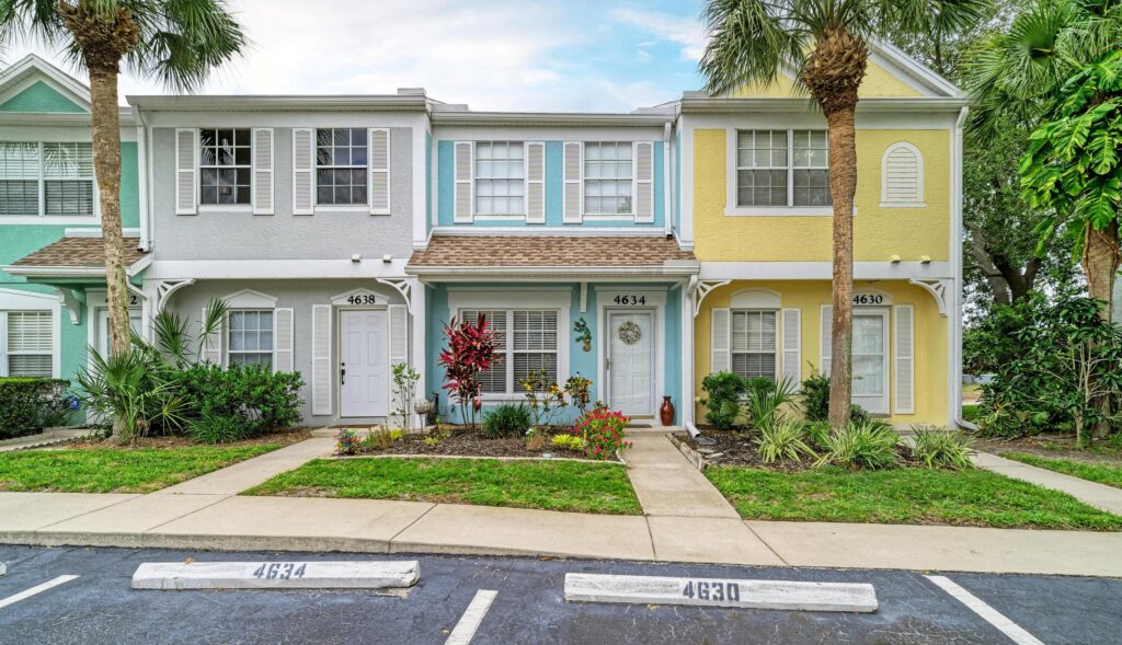 A row of pastel-colored townhouses with palm trees, small gardens, and a parking lot with labeled spaces in front basks in the vibrant energy of leo season. Each house has white trim and a welcoming front entrance, radiating warmth and style under the sunny skies.