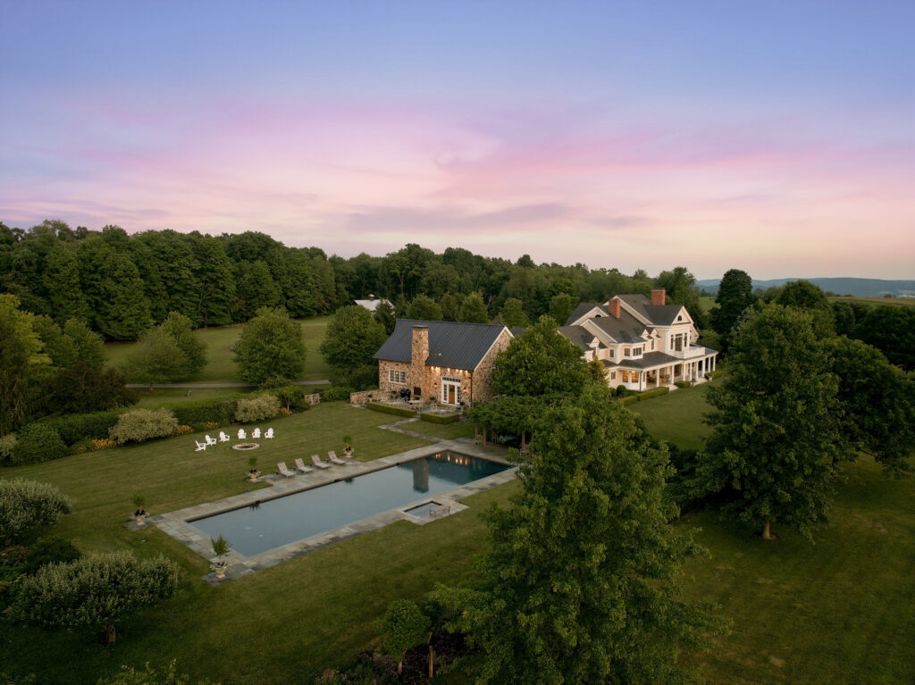 A large country house with a stone exterior, a pool, lounge chairs, and Adirondack chairs on a spacious green lawn, surrounded by trees glowing in the golden light of sunset—perfect for soaking up the vibrant energy of leo season.