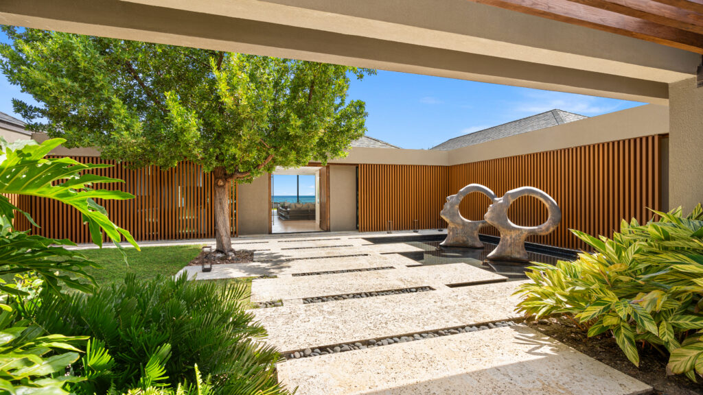 A modern courtyard with a stone path, green plants, two large abstract sculptures, a tree, and wooden slat walls. Designed with summer amenities in mind, the space features an open doorway in the distance offering a refreshing view of the ocean and blue sky.