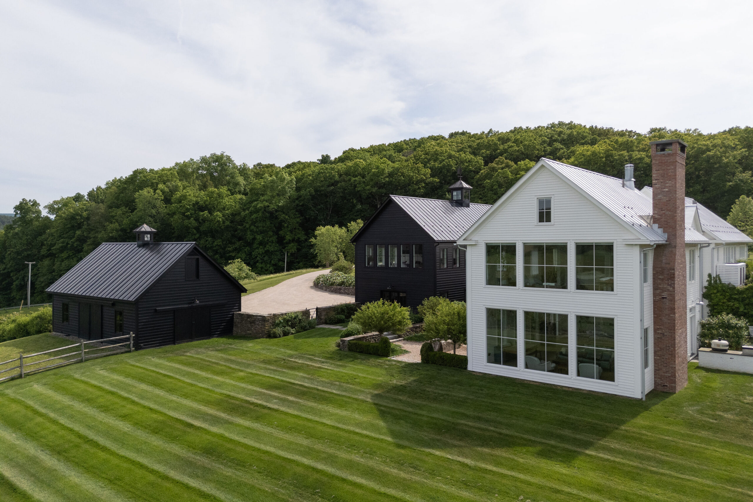 A modern farmhouse with white siding and large windows sits on a neatly mowed lawn, ready to welcome guests for an A-list: August 2026 event, with two black barns nearby and a background of green trees under a cloudy sky.