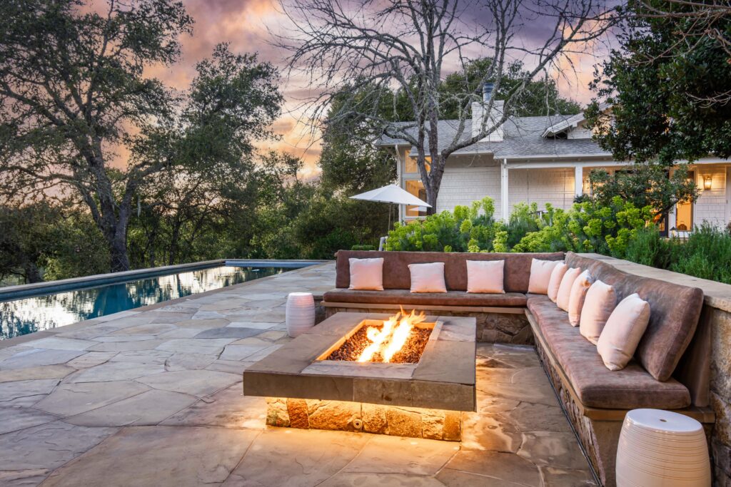 A stone patio with a fire pit, surrounded by a sectional sofa with cushions, sits beside a pool and trees at sunset, with a house and umbrella in the background—an inviting space featuring summer amenities perfect for relaxing or entertaining guests.