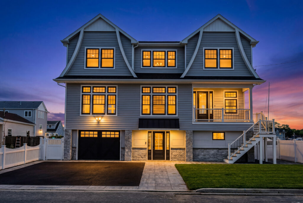 Three-story house at dusk with large lit windows glowing warmly like it's celebrating leo season, gray siding, black garage door, stone accents, covered porch, white fence, green lawn, and stairs leading to the front entrance.