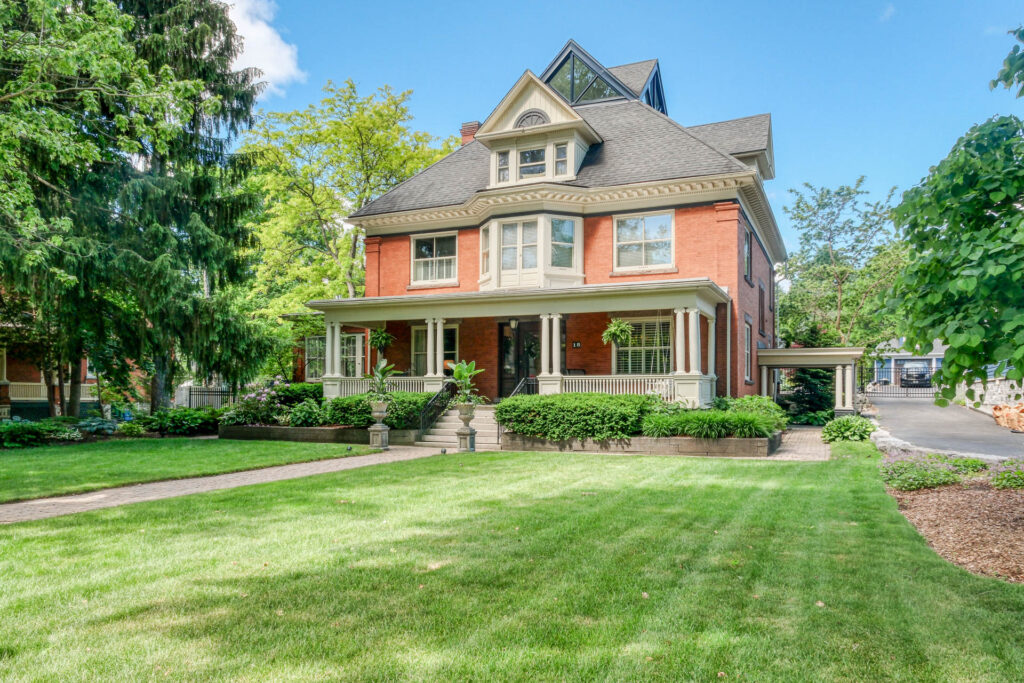 Large red-brick Victorian house with white trim, gabled roof, covered porch, manicured lawn, shrubs, and trees basking under a bright blue sky that captures the vibrant energy of leo season.