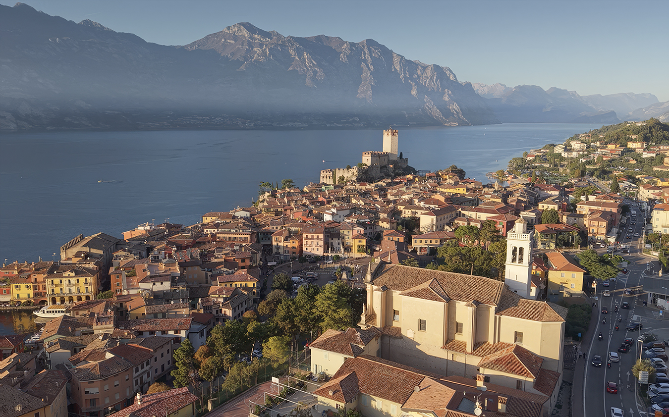 A scenic view of a lakeside town with red-tiled roofs, a castle on a hill, mountains in the background, and calm water reflecting the early morning light.