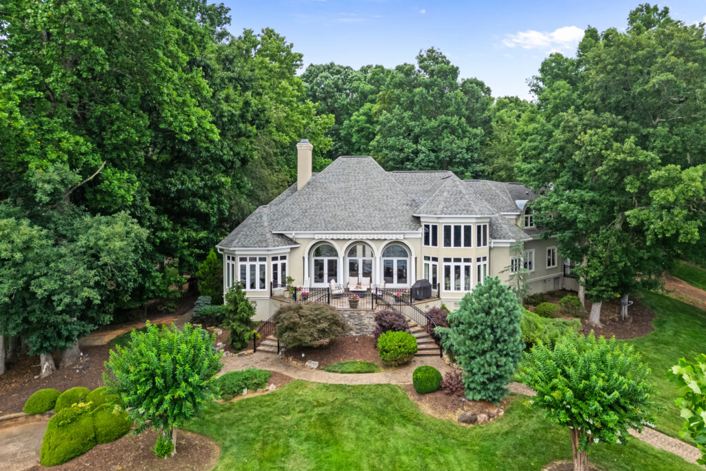 Large two-story house with gray roof and arched windows, surrounded by lush green trees and a landscaped garden, basking in the vibrant energy of leo season, viewed from above on a sunny day.