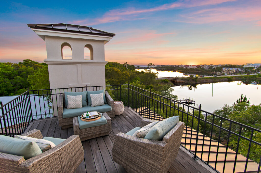 A rooftop patio with wicker chairs, a small table, and cushions overlooks a river and lush trees at sunset, offering summer amenities and a view of distant buildings under a colorful sky.