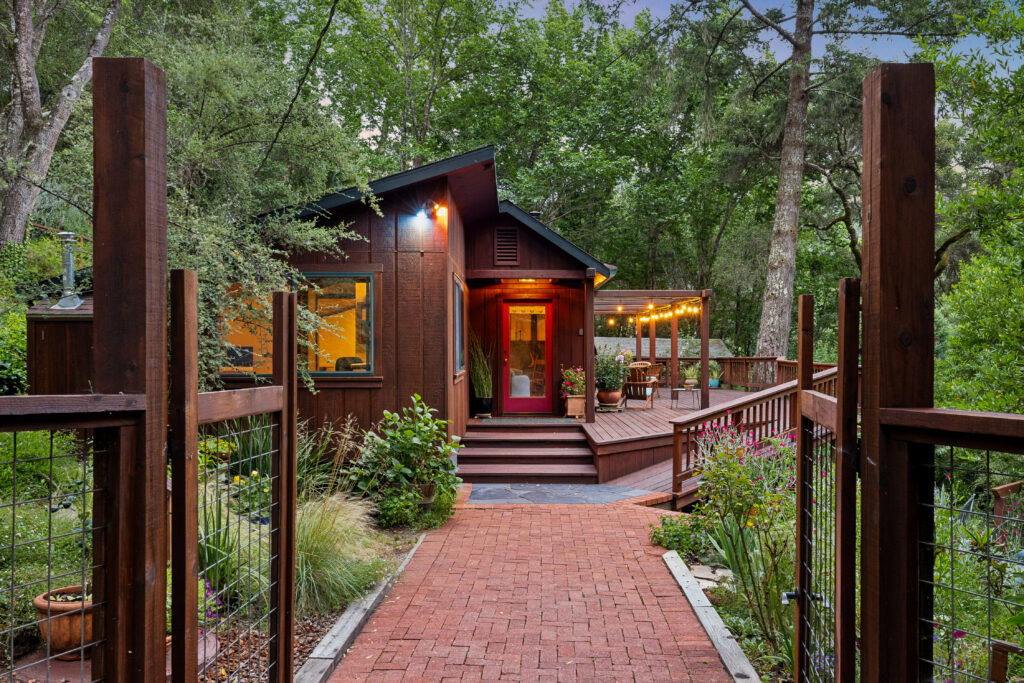 A cozy wooden cabin with large windows and a deck, surrounded by trees and garden, viewed from a brick walkway with a wooden railing and string lights on the patio—perfect for enjoying warm evenings during leo season.