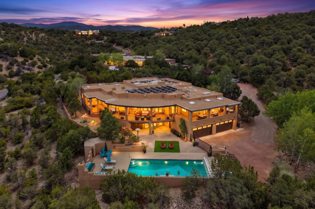 A modern adobe-style house with large windows and a pool, nestled among green hills and trees at sunset during Leo season, with distant mountains under a colorful sky.