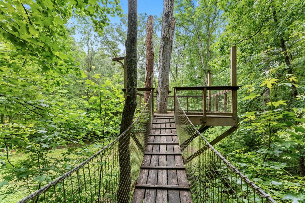 A wooden rope bridge leads to a treehouse platform surrounded by lush green trees in a dense forest under a clear blue sky, offering unique summer amenities for an unforgettable outdoor experience.