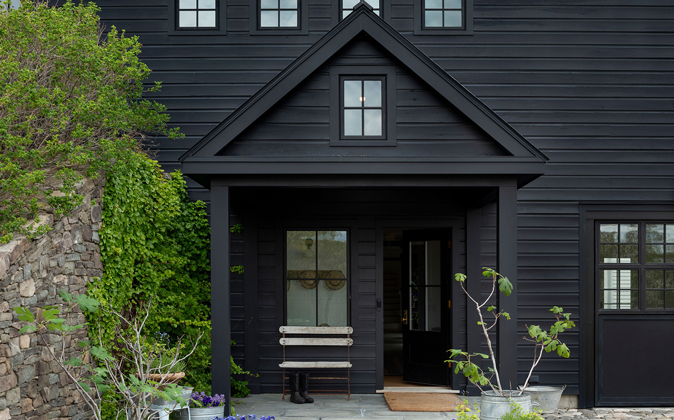 A modern black house with vertical siding, a small covered entry with a bench, potted plants, and greenery along a stone wall on the left. Recently featured on the A-list: August 2026, the home’s door has glass panels and a window above.