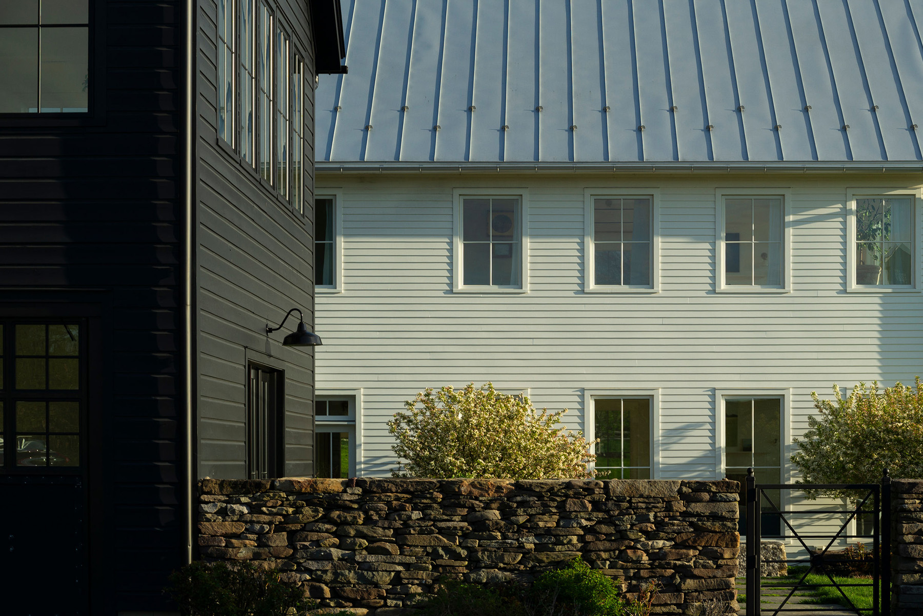 A stone wall separates a dark house on the left and a white house with a metal roof and several windows, with bushes in front and sunlight casting soft shadows—a tranquil August 2026 afternoon scene.