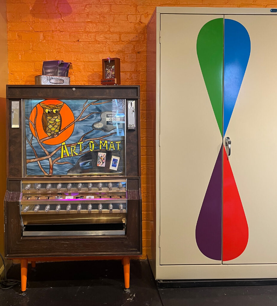 A retro Art-O-Mat vending machine with an owl design stands next to a beige cabinet decorated with colorful teardrop shapes, against an orange brick wall.