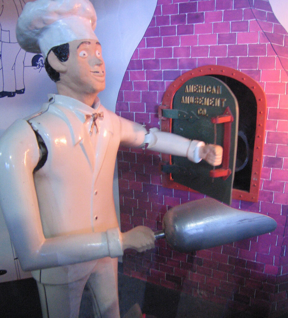 A chef mannequin in white attire holds a pizza peel in front of a brick oven with an open door labeled AMERICAN AMUSEMENT. The scene appears to be part of an animated display or diorama.