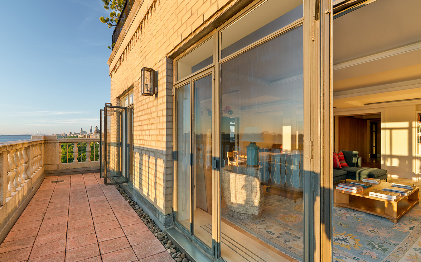 Sunny balcony with a tiled floor and glass doors leading to a cozy living room with a rug, coffee tables, and books; city skyline is visible in the distance.