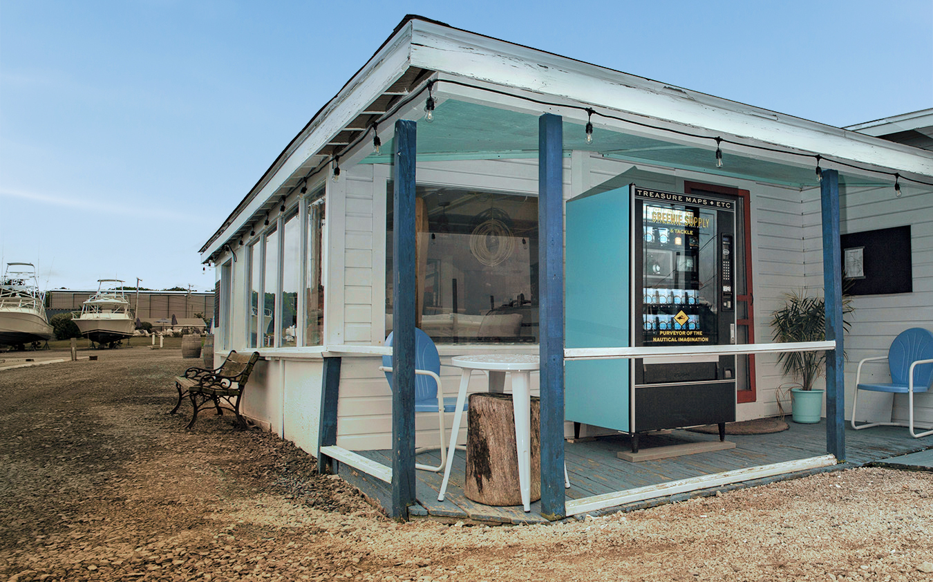 A vending machine stands on the porch of a small white building with blue trim. There are two blue chairs, a small table, and boats visible in the background. The area is rustic with gravel on the ground.