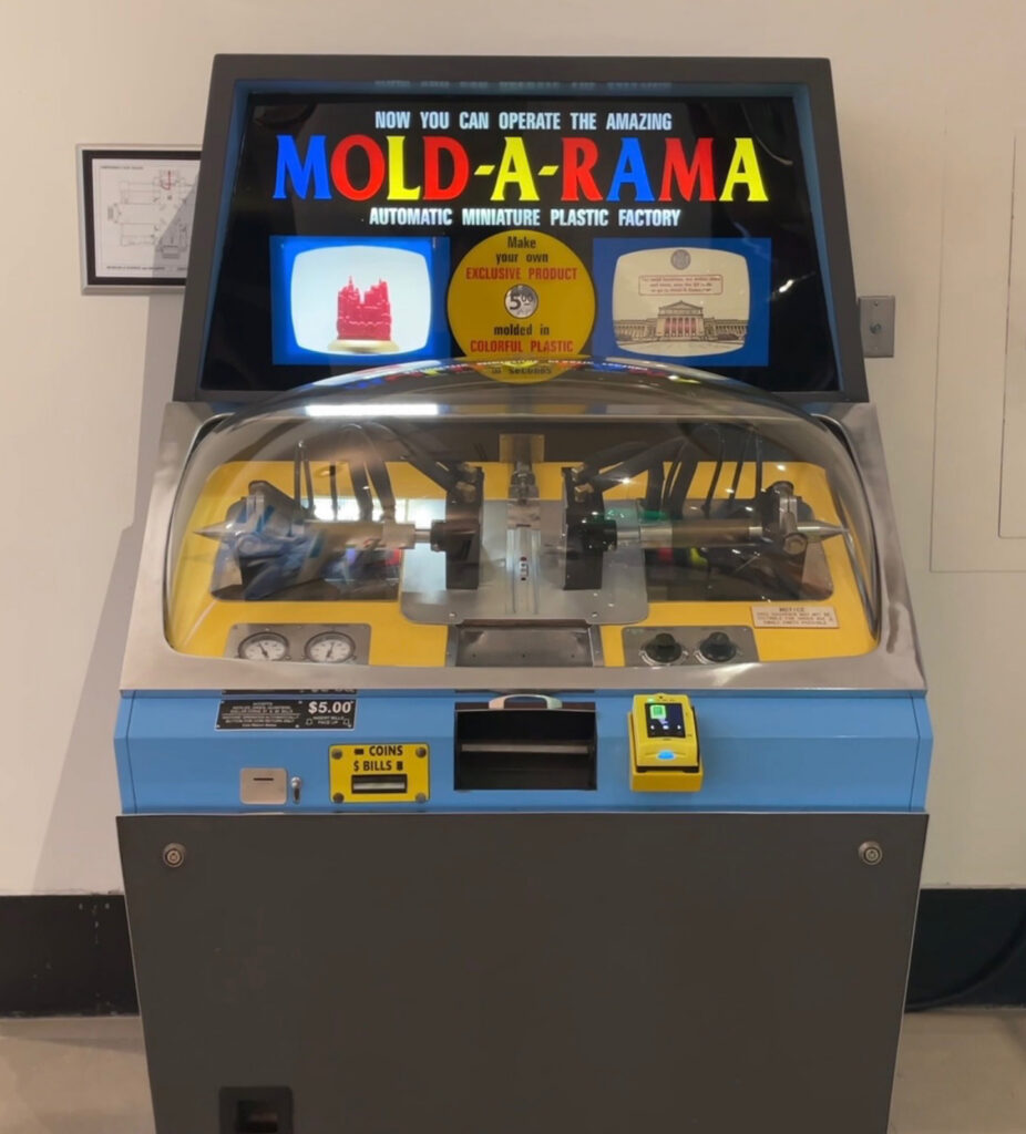 A vintage Mold-A-Rama machine with a glass dome, colorful display, and payment slots. The sign advertises making plastic souvenirs for $5.00, showing example molds of a red alligator and a building.