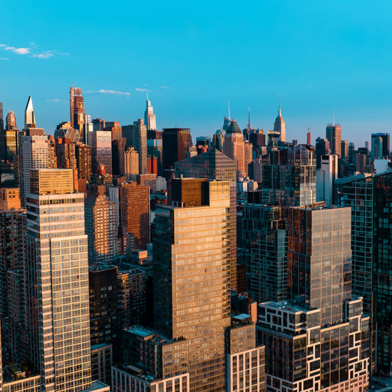 Aerial view of New York City skyscrapers at sunset, with sunlight reflecting off the glass buildings and a clear blue sky above—a stunning backdrop that highlights the vibrant world of NYC residential rental living amid iconic city sights.