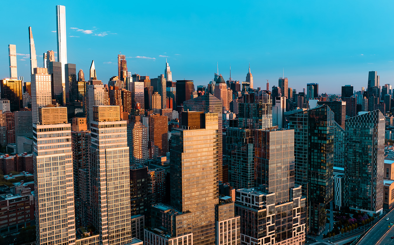 Aerial view of New York City skyscrapers at sunset, with sunlight reflecting off the glass buildings and a clear blue sky above—a stunning backdrop that highlights the vibrant world of NYC residential rental living amid iconic city sights.