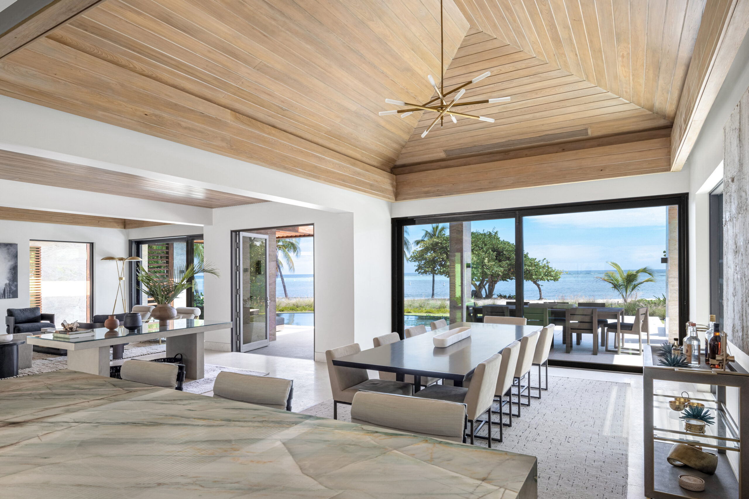 Modern open-concept dining and living area with wood ceiling, long dining table, large windows, and a view of palm trees and the ocean outside. Light neutral tones and contemporary decor create an inviting space perfect for hosting an a-list August 2026 gathering.
