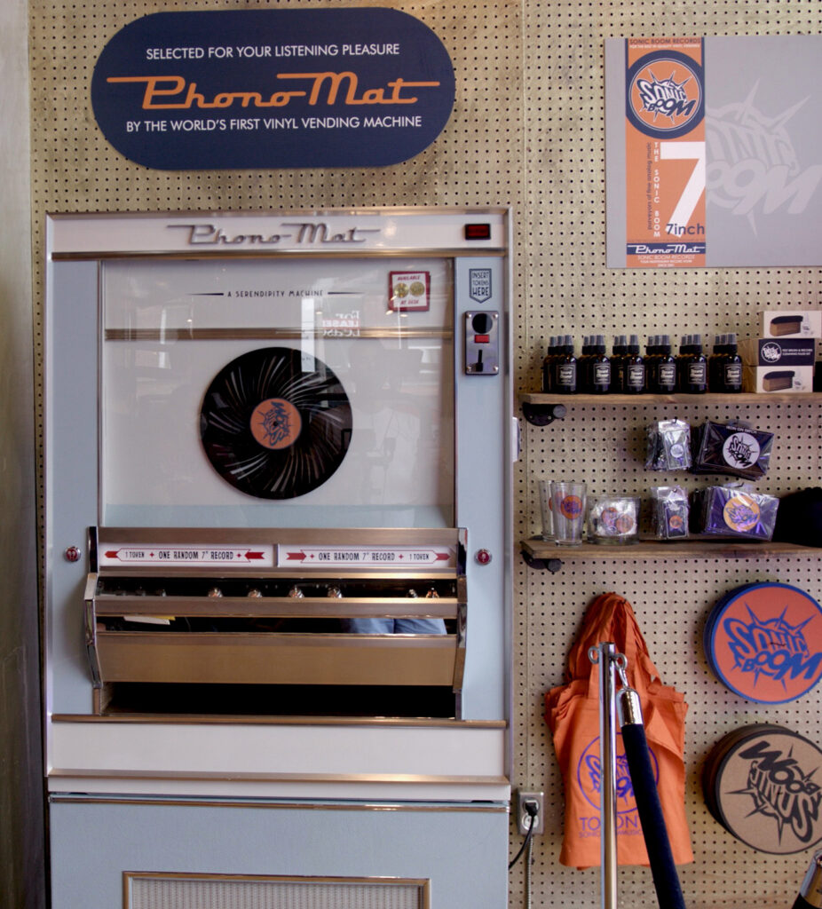 A retro vinyl vending machine labeled “PhonoMat” with records, mugs, and accessories displayed on a pegboard wall nearby. A large vinyl record and branded merchandise are also visible.