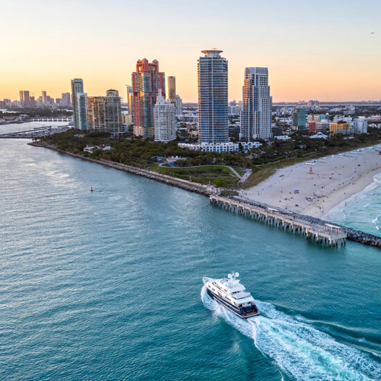 Aerial view of a yacht sailing near a beach with high-rise buildings, a sandy shoreline, and the ocean at sunset in a coastal city—an iconic scene that captures the vibrant allure of South Florida Real Estate.