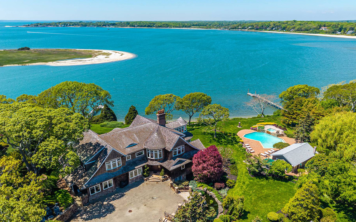 A large waterfront mansion with a circular driveway, surrounded by trees, a swimming pool, and a pool house. A long dock extends into a blue bay, with a sandy beach and greenery in the background.
