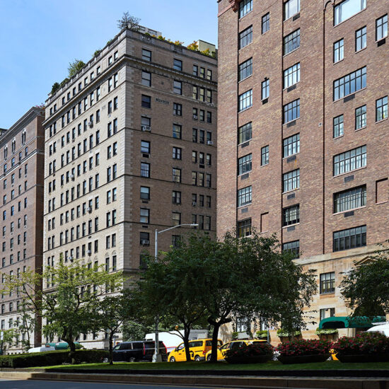 A row of tall brick apartment buildings lines a city street in Manhattan, with trees, flowerbeds, and yellow taxis parked along the curb—an inviting scene reflecting the area’s thriving luxury sales under a clear blue sky.