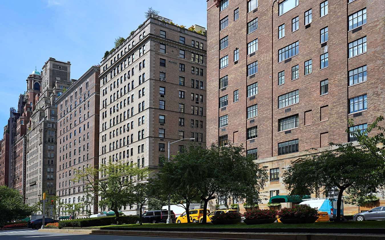A row of tall brick apartment buildings lines a city street in Manhattan, with trees, flowerbeds, and yellow taxis parked along the curb—an inviting scene reflecting the area’s thriving luxury sales under a clear blue sky.
