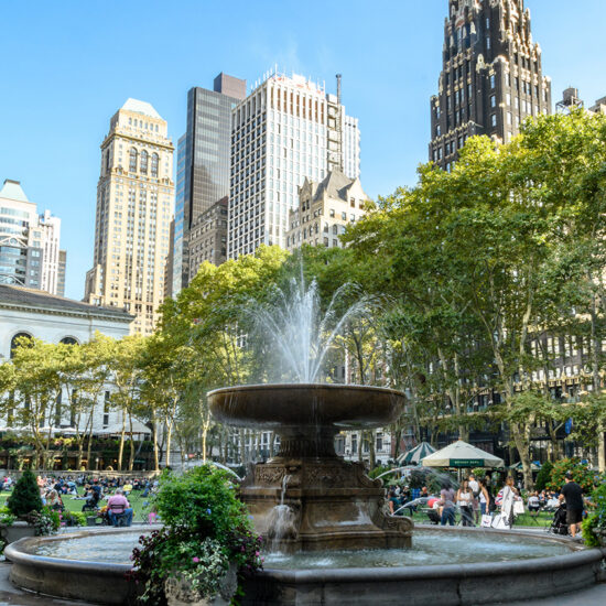 A large fountain in a city park with people sitting and relaxing, set against the backdrop of tall skyscrapers and leafy green trees. Nearby, elegant Manhattan condos rise above the scene under a clear blue sky.