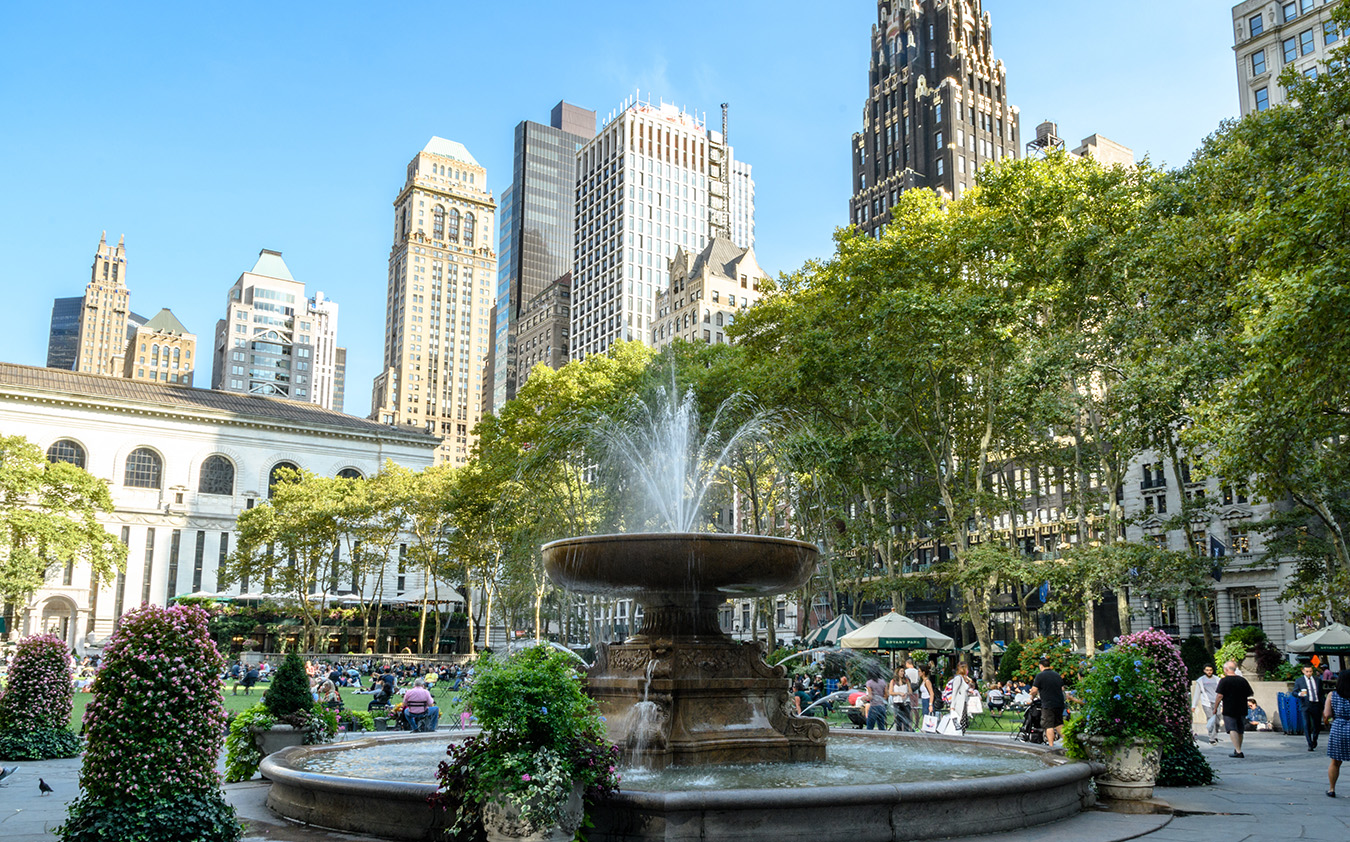 A large fountain in a city park with people sitting and relaxing, set against the backdrop of tall skyscrapers and leafy green trees. Nearby, elegant Manhattan condos rise above the scene under a clear blue sky.