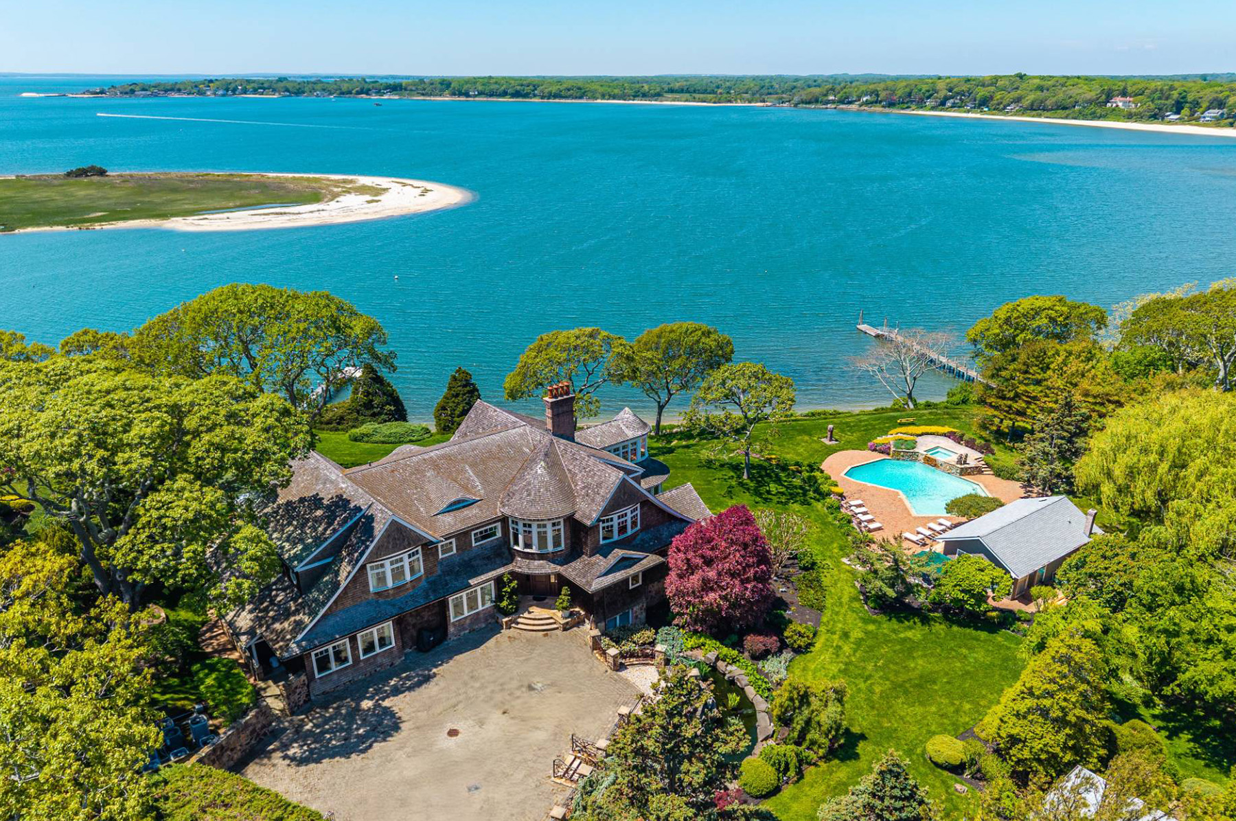 A large waterfront estate with a stone mansion, pool, and landscaped gardens, surrounded by trees, overlooking a blue bay with a sandy shoreline and distant land across the water.