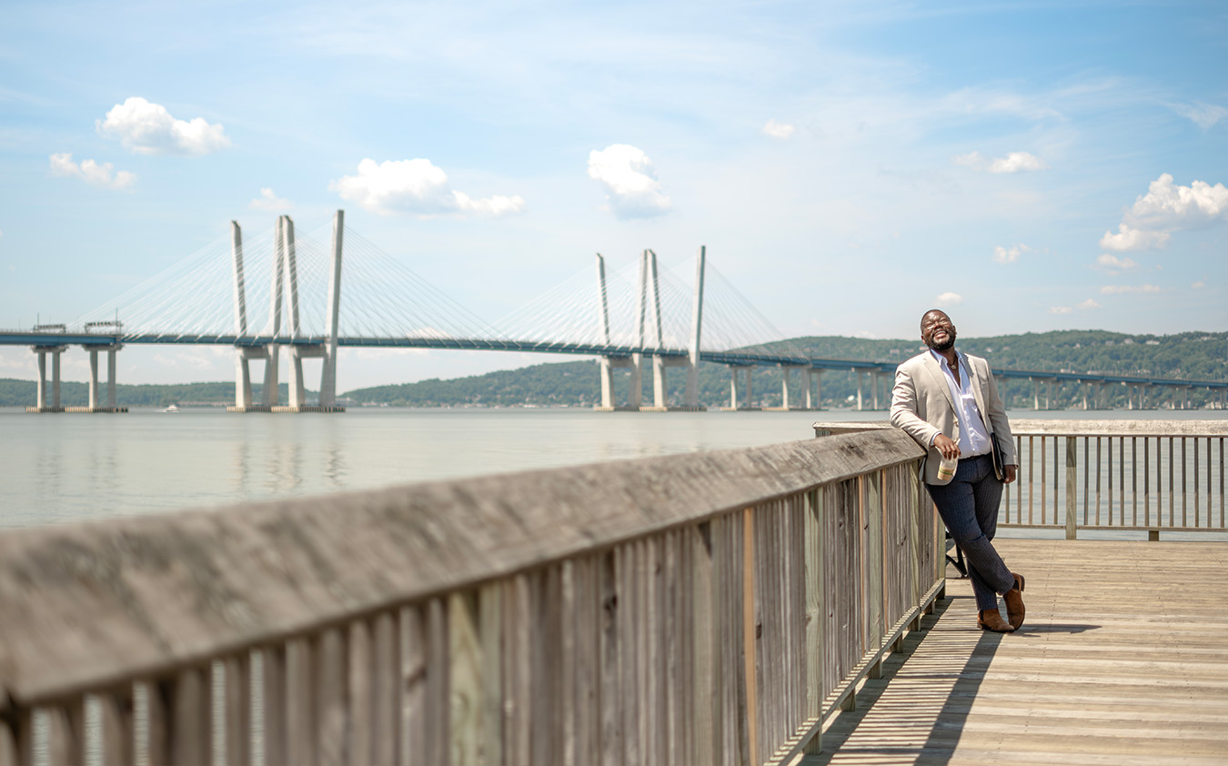 A person in a light blazer leans against a wooden railing on a pier, smiling, with a large cable-stayed bridge and calm water in the background under a blue sky with scattered clouds.