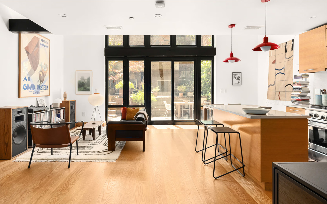 Modern living room and kitchen with wood floors, large windows, bar stools, leather chairs, art on the walls, and a view of a green outdoor space. Bright, airy, and stylish open-concept space.