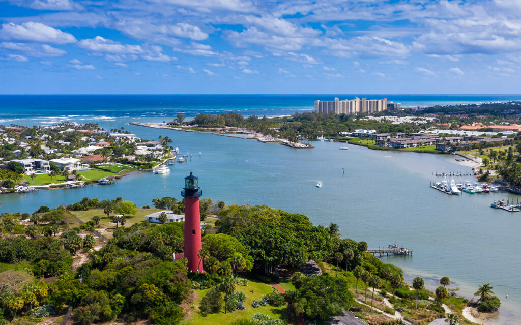 Aerial view of a coastal town with a red lighthouse, river, marina, waterfront homes, lush greenery, blue ocean, and a cluster of tall buildings in the distance under a partly cloudy sky—a stunning snapshot that captures the essence of South Florida Real Estate.