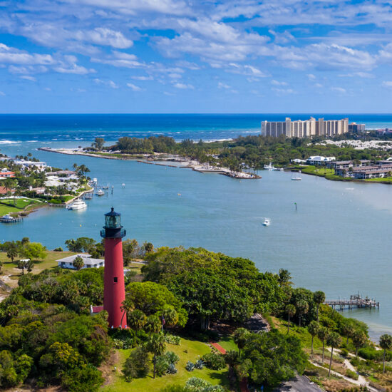 Aerial view of a coastal town with a red lighthouse, river, marina, waterfront homes, lush greenery, blue ocean, and a cluster of tall buildings in the distance under a partly cloudy sky—a stunning snapshot that captures the essence of South Florida Real Estate.